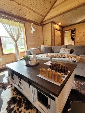 A cozy cabin interior with a wooden ceiling, a fireplace, a sofa, and a chessboard. - Bohío Retreat - Cabin and Creek View on 42 Acres (Steelville)