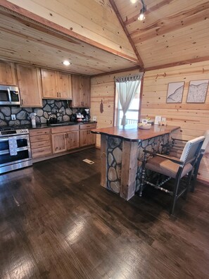 A kitchen with wooden cabinets, a stone island, and a dining chair. - Bohío Retreat - Cabin and Creek View on 42 Acres (Steelville)