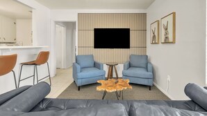 Ein modernes Wohnzimmer mit Flachbildfernseher, zwei blauen Sesseln, einem Holztisch und einem grauen Ledersofa.