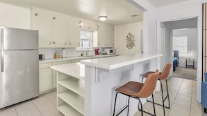 Eine moderne Küche mit weißen Schränken, einem Edelstahl-Kühlschrank, einer großen Kücheninsel mit Barhocker und Blick in das angrenzende Schlafzimmer.