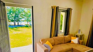 A living room with a beige sectional sofa, a small side table, and a window with curtains. - Cozy, comfortable 1-bedroom apartment in charming Eleuthera near to the beach. (North Eleuthera)