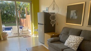 Ein Wohnzimmer mit grauem Sofa, einem Holztisch, einem weißen Kühlschrank und einem großen Fenster mit Blick auf einen Pool und Grünflächen.