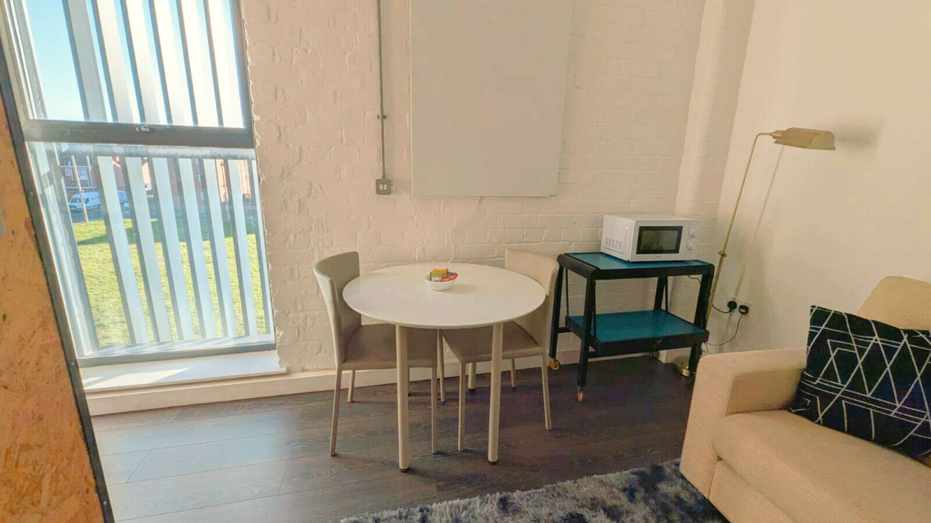 A small, well-lit room with a white table, two chairs, a microwave on a small table, a sofa, and a view through a window.