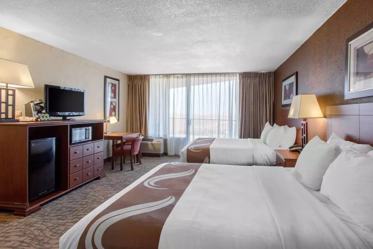 A hotel room with two beds, a desk with a TV, a minibar, and a large window with curtains.