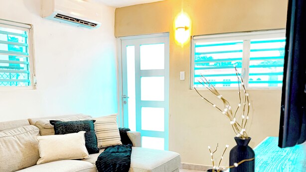 Een woonkamer met een beige bank, een houten salontafel en een televisie. Er zijn twee ramen met zonwering, een wandairconditioner en een wandlamp.