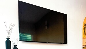 Een moderne woonkamer met een flatscreen televisie aan de muur, een houten kast met etalageglas deuren, en twee donkere vazen met takken.
