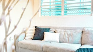 Een woonkamer met een beige bank, een decoratief plaid en een raam met zonwering.
