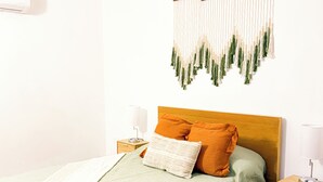 Een houten bed met een groene sprei en twee oranje kussens, een nachtkastje met een lamp, en een wanddecoratie van macramé.