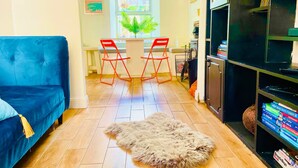 Een woonkamer met een blauwe bank, een kleedje, een eettafel met rode stoelen en een boekenplank.