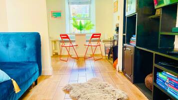 A living room with a blue sofa, a small rug, a dining table with red chairs, and a bookshelf.