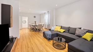 A modern living room with a sofa, coffee table, and TV. There is a dining area with chairs and a table in the background.