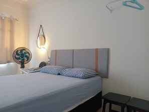 A bed with a grey headboard, a bedside table with a lamp, a fan, and a bench. - Capsula Residence Hotel (Marília)