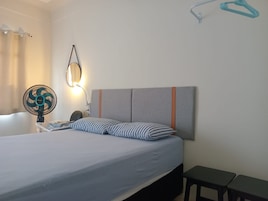 Uma cama com cabeceira cinza, mesa de cabeceira com abajur, ventilador e um banco.