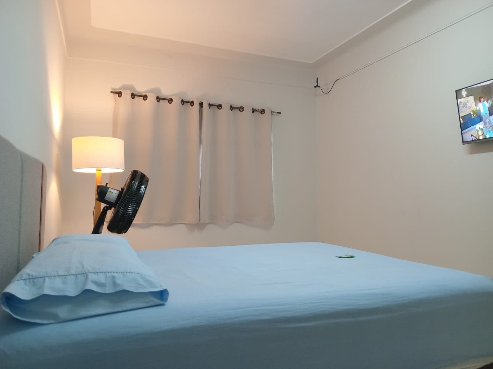 Et hotelværelse med en seng, en ventilator, en lampe og et vindue med gardiner.
