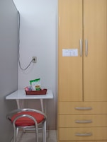 Um quarto pequeno com uma mesa branca, uma cadeira vermelha, um guarda-roupa de madeira com gavetas e uma tomada.