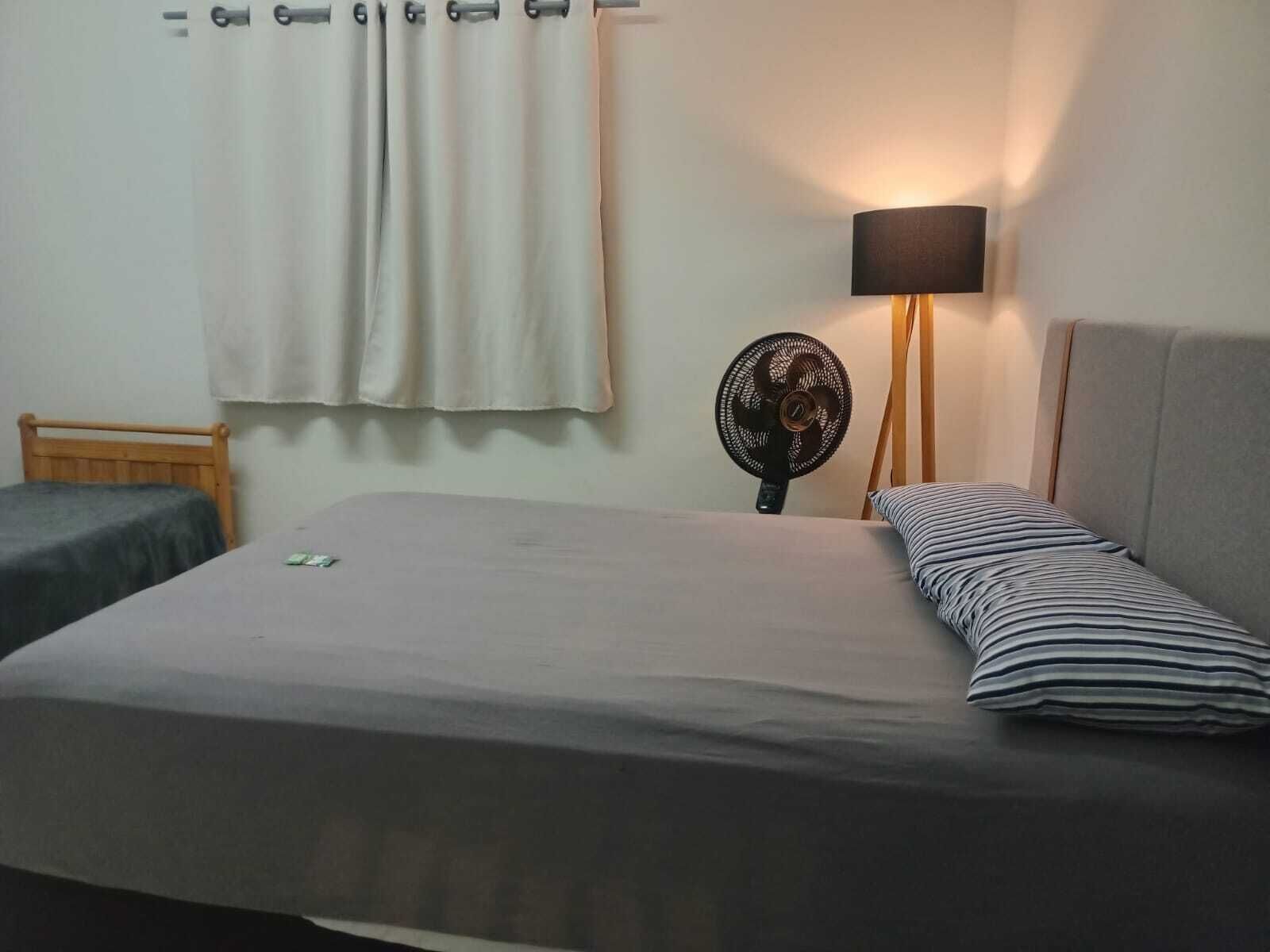 Una cama individual con colchón gris y almohada a rayas, una mesita de noche con lámpara, y una ventana con cortinas.