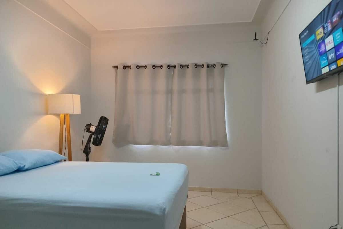 A bedroom with a bed, a lamp, a guitar, a TV, and a window with curtains.