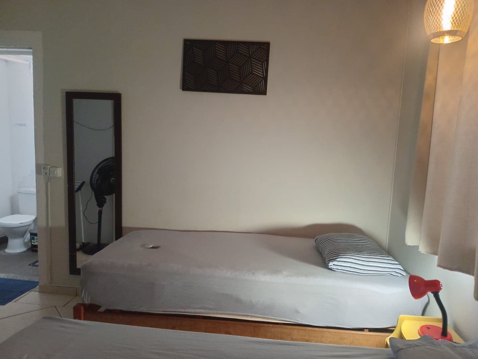 Ein Einzelbett mit Kissen, ein Nachttisch mit Lampe, ein Spiegel und eine Wandleuchte.