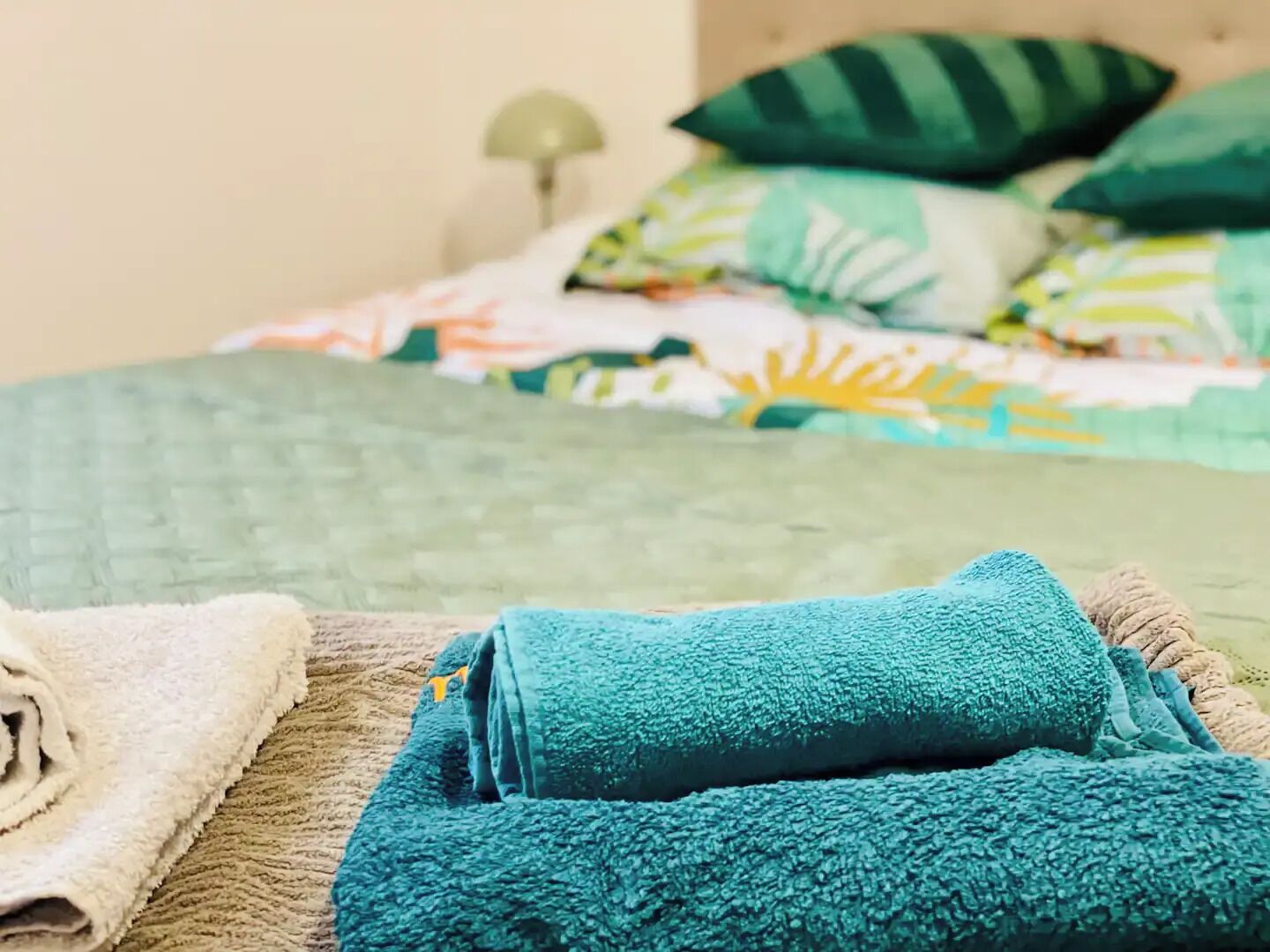 A neatly made bed with a green quilt and multiple pillows, a rolled white towel, and a beige blanket on a textured surface.