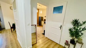 Un pasillo con suelo de madera, paredes blancas y una puerta que da a una zona de cocina. Hay plantas en macetas sobre una pequeña mesa cerca de la puerta.