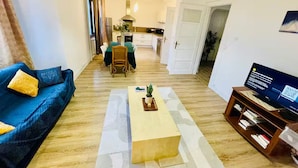 Una sala de estar con un sofá, una mesa de centro, un mueble para la televisión y vista hacia la cocina.