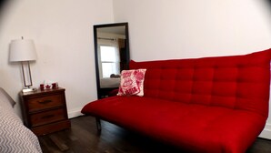 Une chambre avec un lit rouge, une table de chevet en bois avec une lampe, un miroir et une fenêtre avec des rideaux.