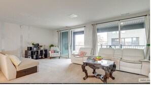 A spacious living room with a beige sofa, a white sofa, and a glass-top coffee table. There is a bookshelf and a chair with colorful cushions. - Delightful balti, Washington DC retreat (Laurel)