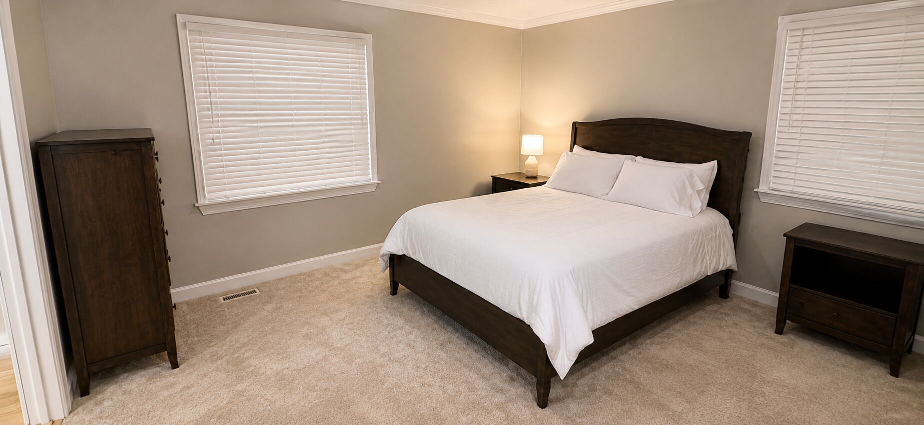 A bedroom with a bed, nightstand, dresser, and two windows with blinds.