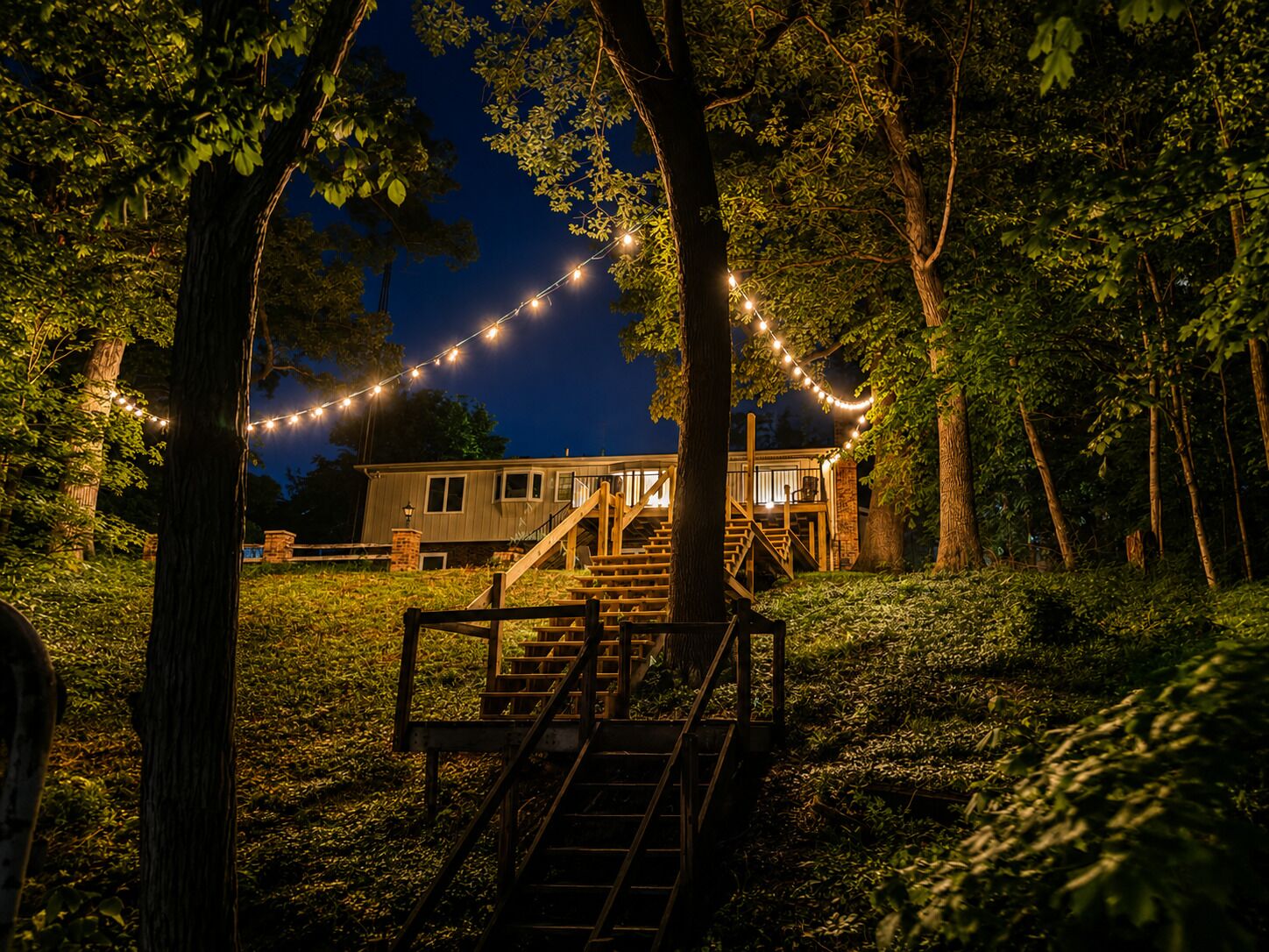 A house with a wooden staircase leading to a deck, surrounded by trees and string lights.