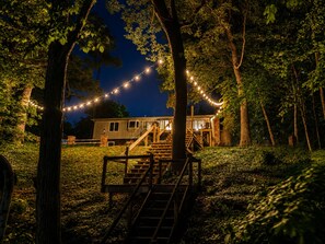 Et hus med en trætrappe op til en terrasse, omgivet af træer og lyskæder.