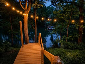 Eine Holzterrasse führt zu einem Steg über dem Wasser, umgeben von Bäumen und Lichterketten.
