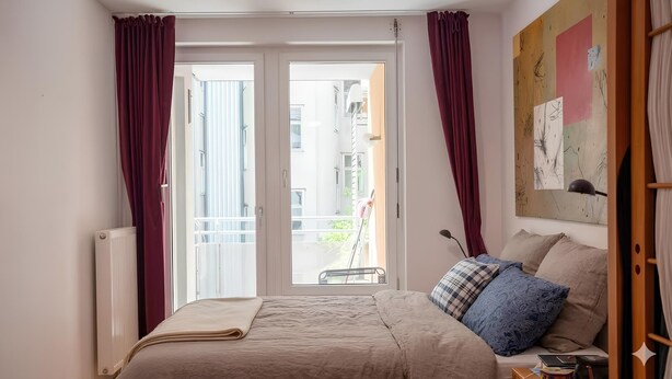 Et soveværelse med seng, radiator, et vindue med gardiner og udsigt til en balkon.