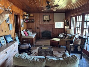 A rustic cabin interior with a fireplace, wooden walls, a sofa, a chair, and a deer head decoration.