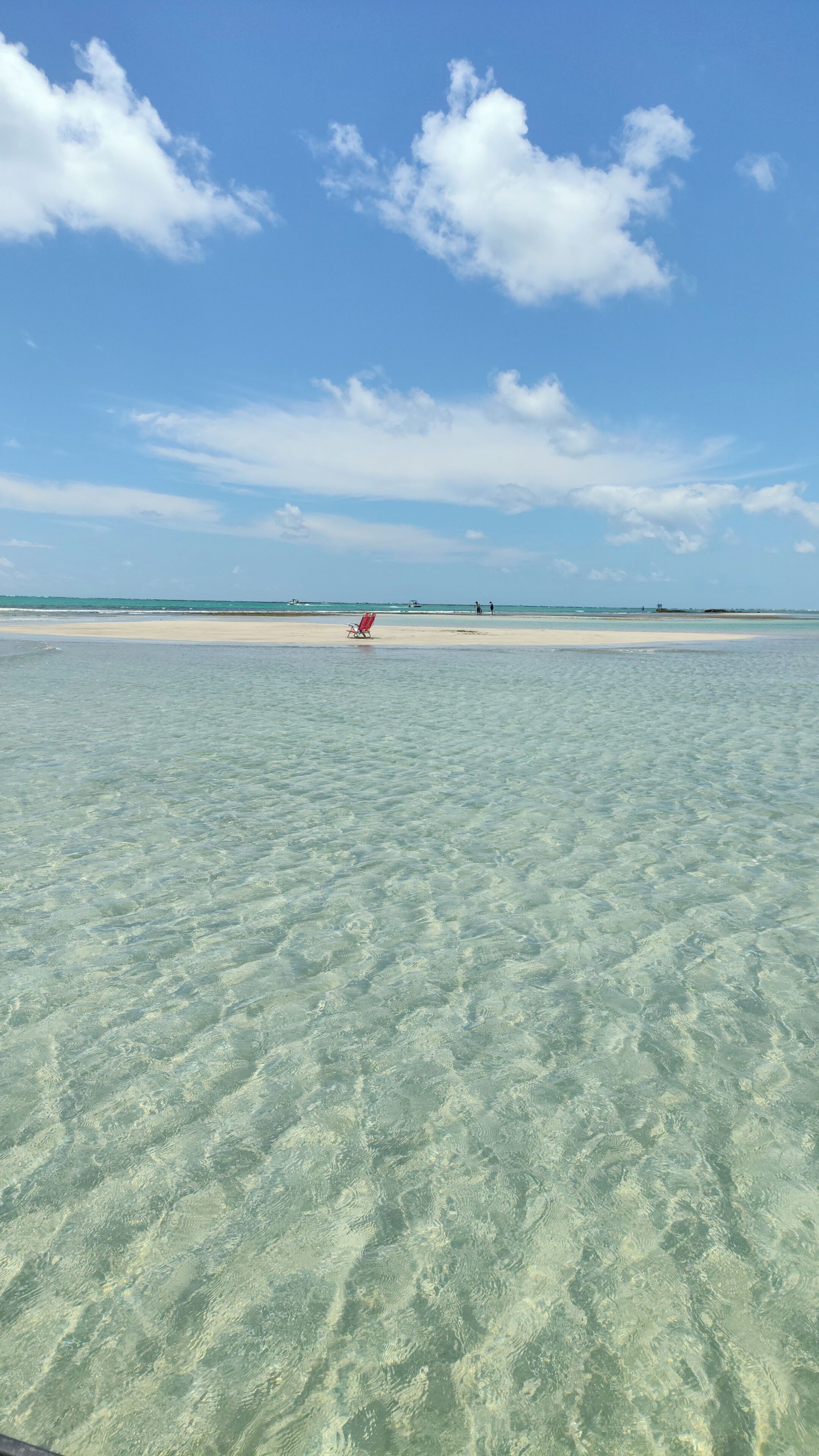 A beach with clear shallow waters, a sandy shore, and a few people in the distance.