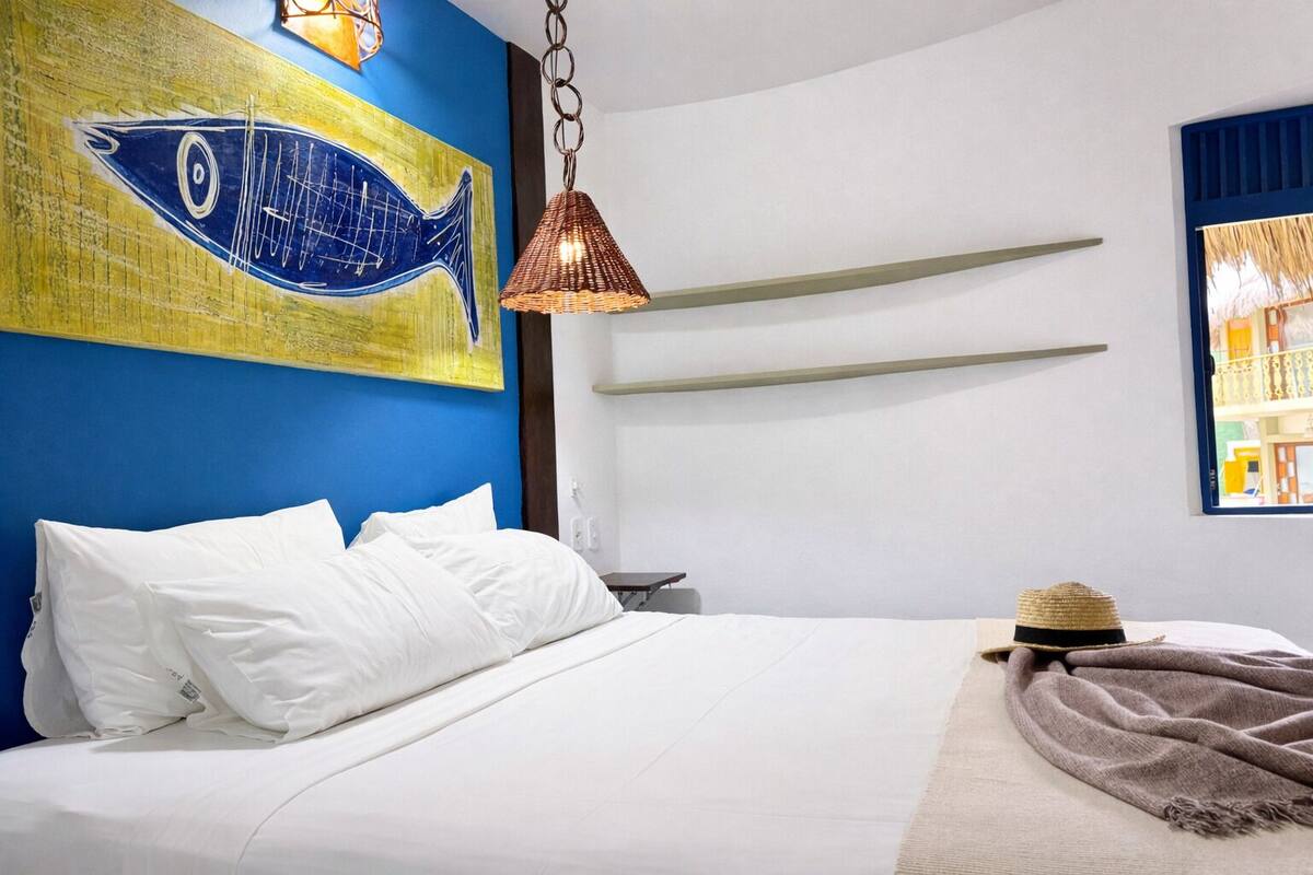 A bedroom with a bed, white pillows, a blue and yellow fish painting, a copper pendant light, and a straw hat on a blanket.
