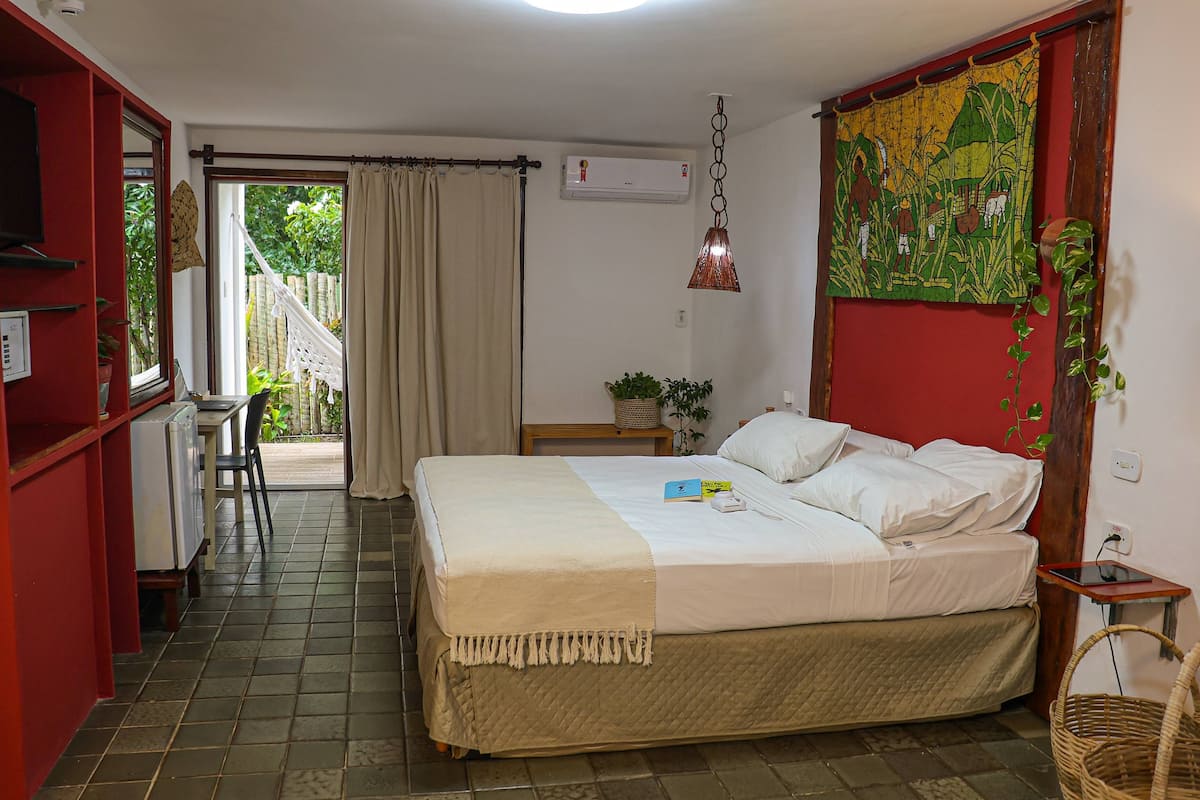 A room with a bed, a desk, a chair, a window with curtains, a hanging plant, and a wall tapestry.