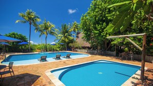 A swimming pool with two sections, surrounded by palm trees and a thatched-roof structure.