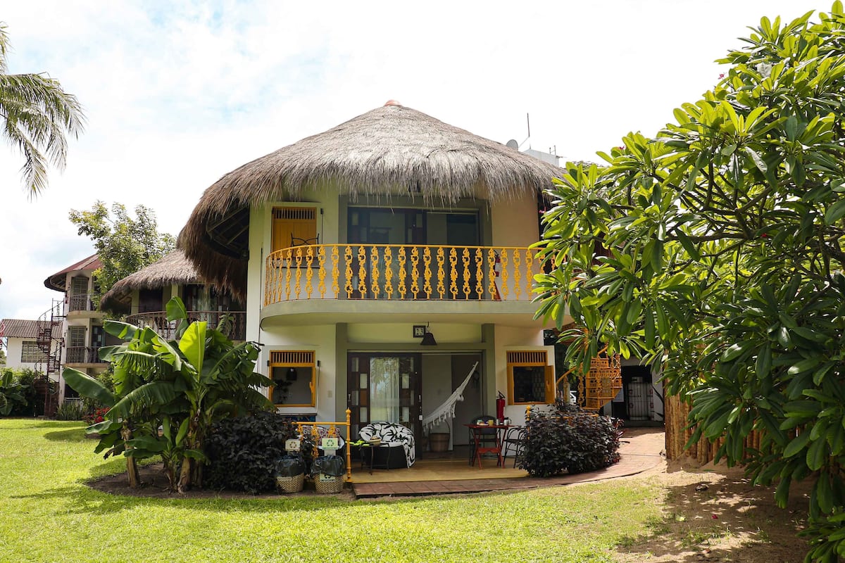 A two-story building with a thatched roof, a balcony with yellow railings, and a well-maintained garden.