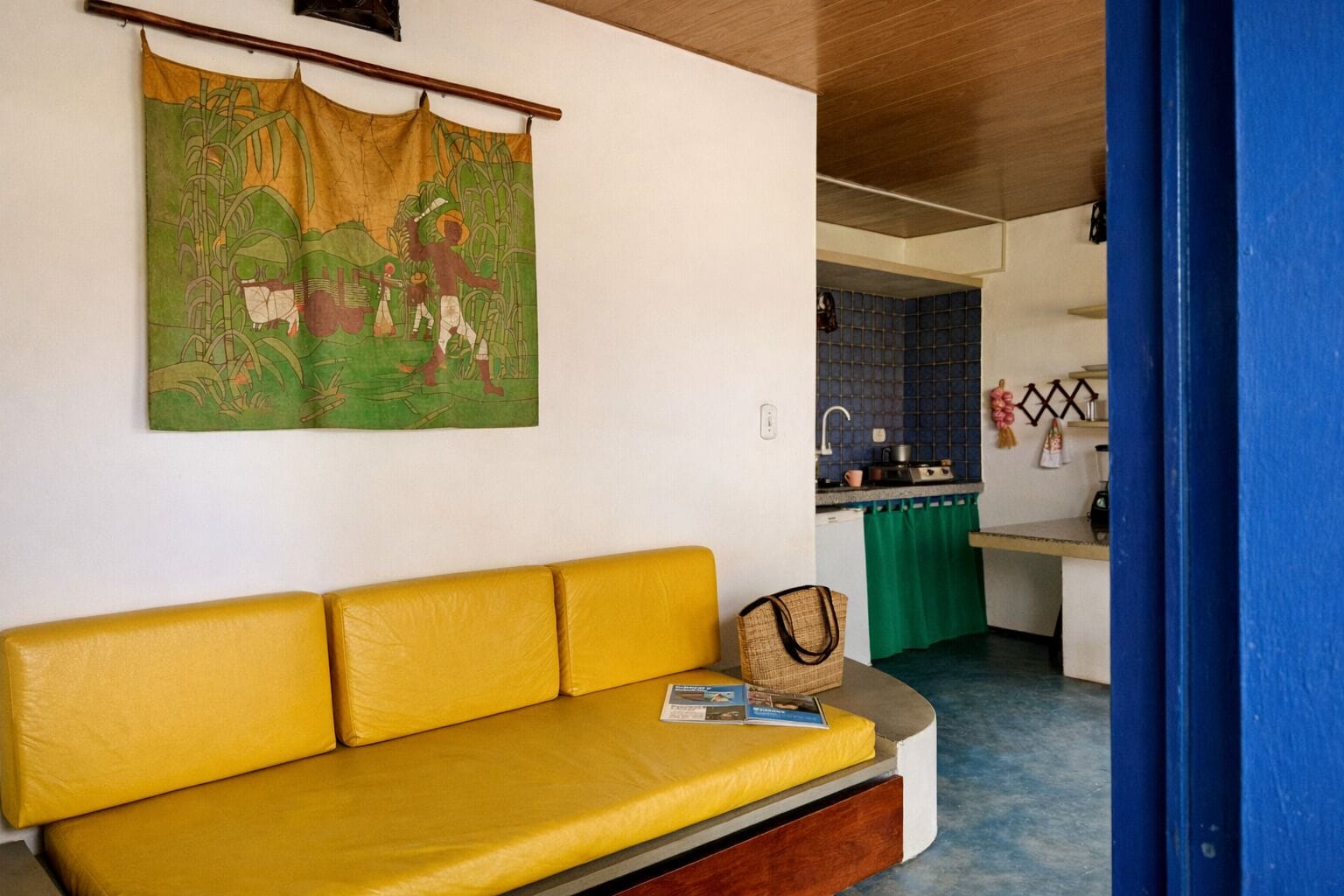 Un sofá amarillo con un bolso tejido y una revista, un tapiz verde con una escena campestre, y una cocina con salpicadero de azulejos azules.