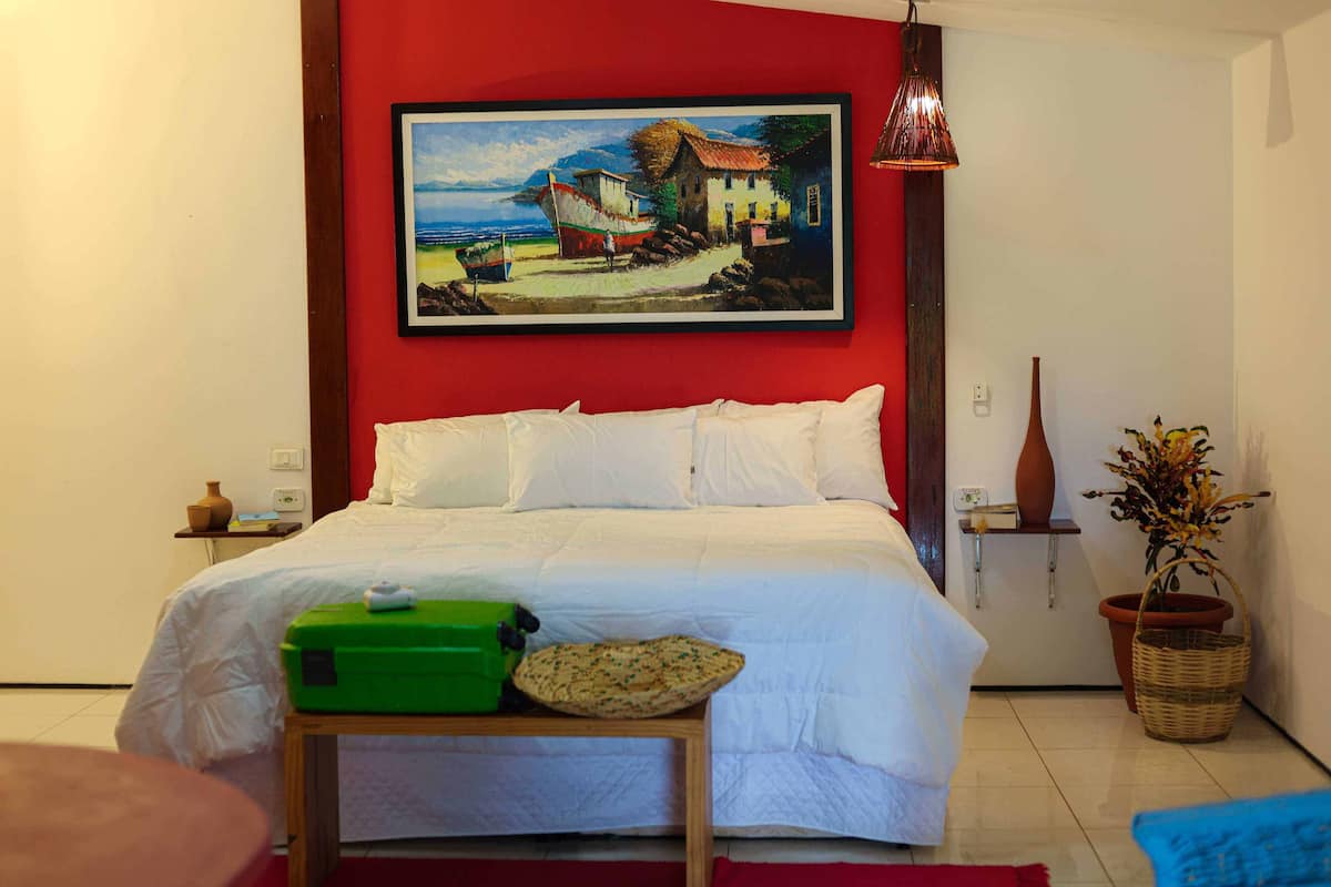 A bedroom with a bed, a painting, a green suitcase, a red rug, and a potted plant.