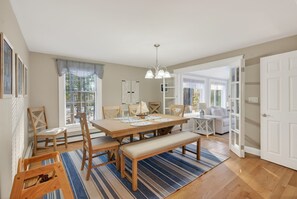 Een eetruimte met een houten tafel, stoelen en een bank. Er is een raam met jaloezieën, een kroonluchter en uitzicht op de omgeving.