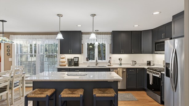 A modern kitchen with dark cabinets, a central island with stools, stainless steel appliances, and pendant lighting. - Walk to Beach&Town! 4 Bedrooms|Riverfront|Pet Friendly|Arcade game (Kennebunk)