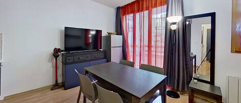 Ein gemütlicher Wohnraum mit Esstisch, Stühlen, Fernseher, Kühlschrank und einem fenster mit Vorhängen.