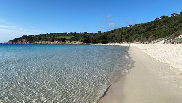 Una playa con aguas transparentes y poco profundas, una orilla arenosa y una colina con vegetación.