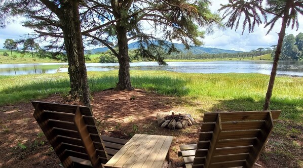 A wooden bench and table set up under trees by a river with a view of a green landscape and hills in the distance. - Ilhas do Perimbó (Otacilio Costa)