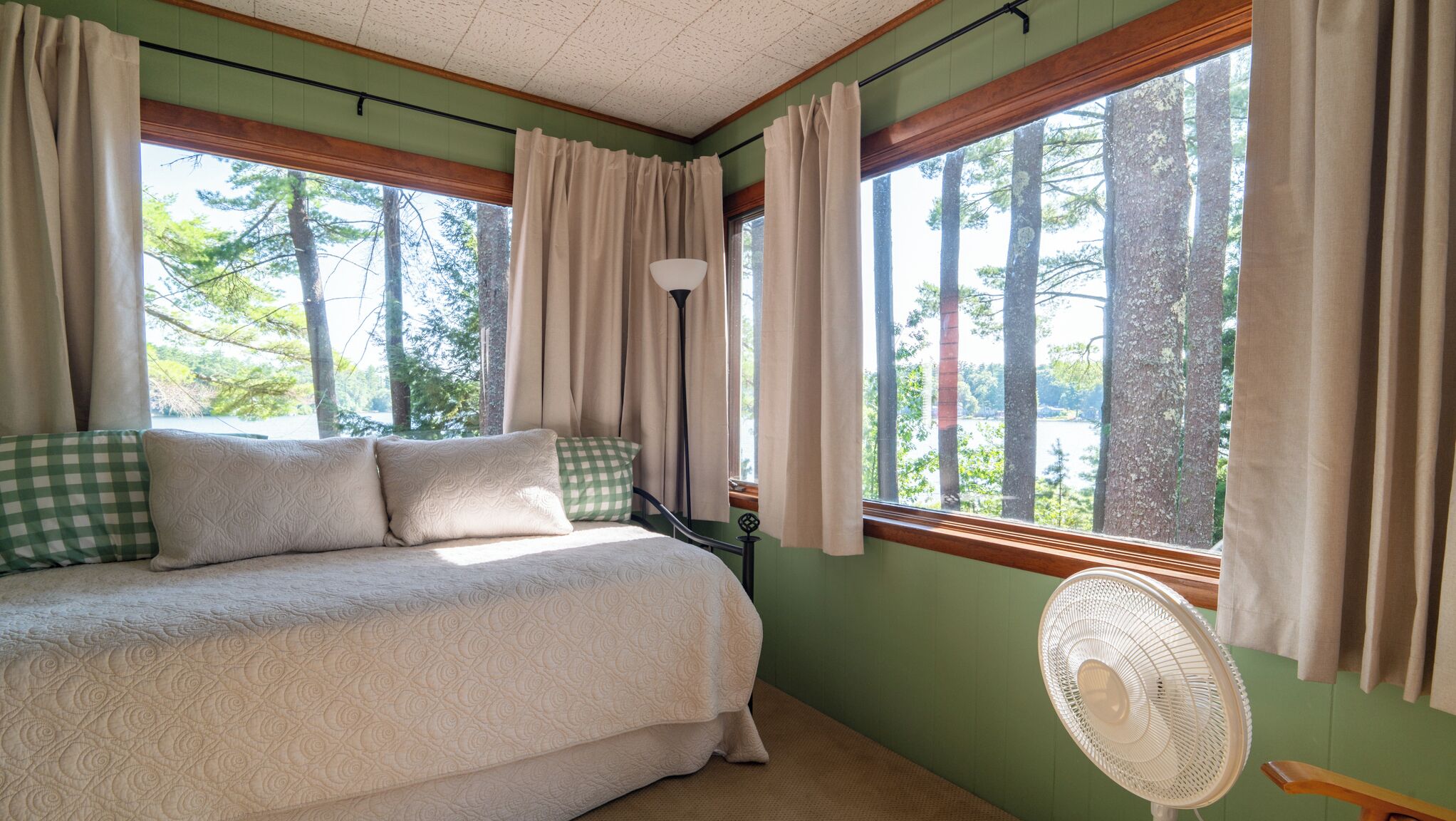 A bedroom with a bed, a standing fan, and large windows with curtains.