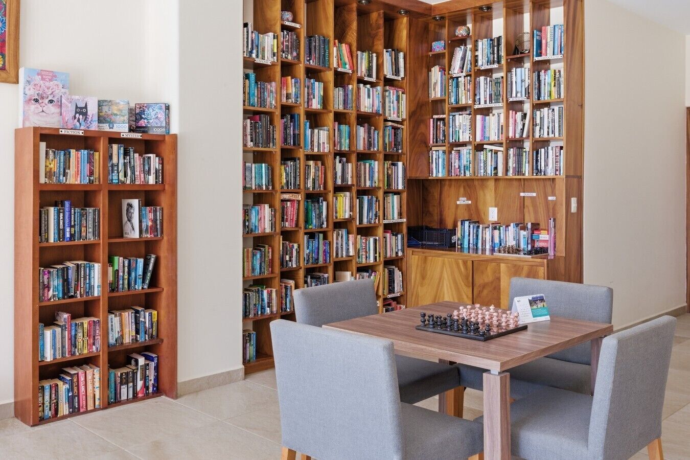A library with multiple wooden bookshelves filled with books, a chess table, and grey chairs.
