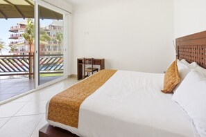 A hotel room with a bed, a wooden headboard, a balcony with a view of buildings and palm trees. - Ocean Front - Two Bedroom Vivo Resorts (Bajos de Chila)