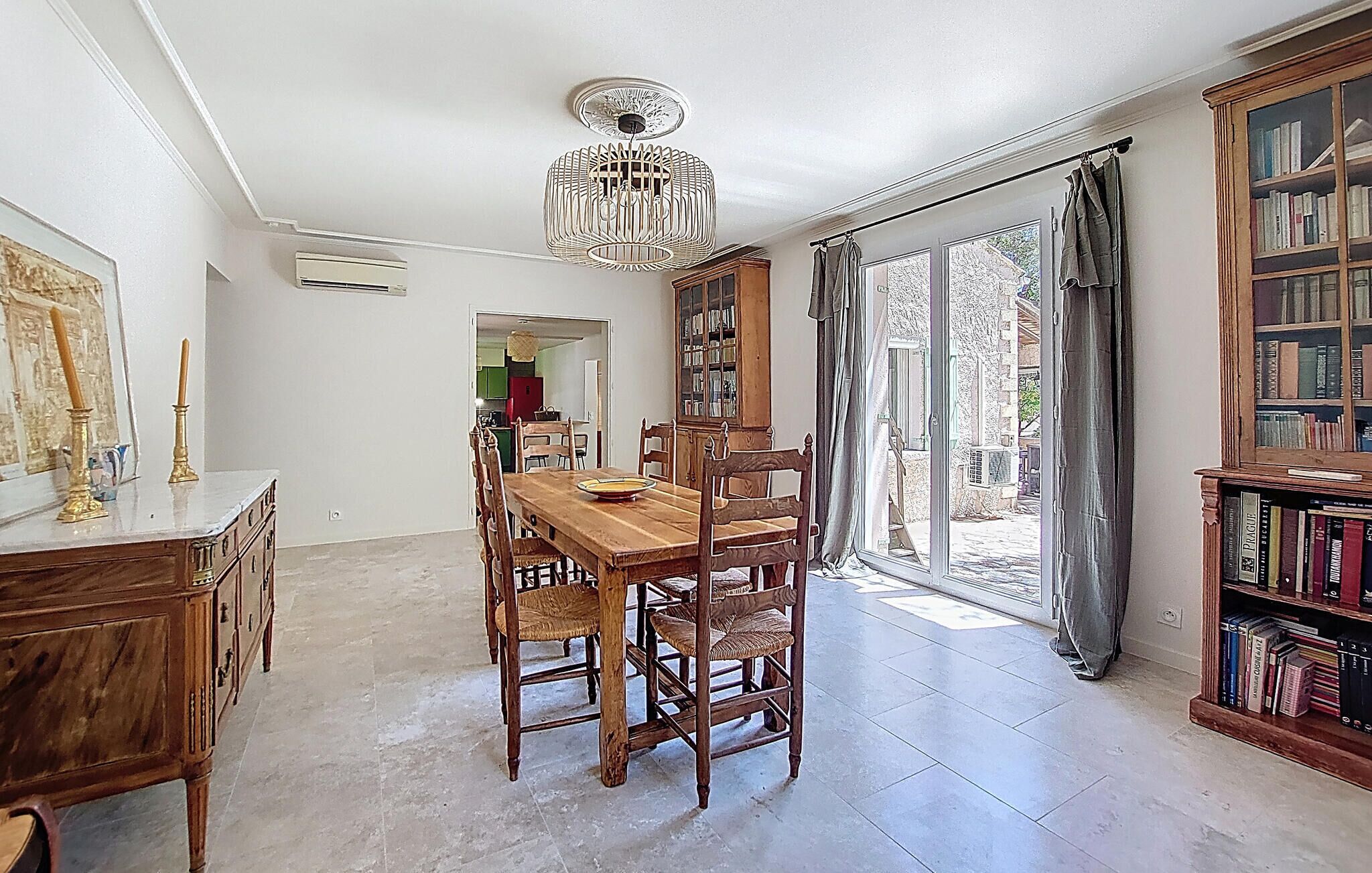 Une salle à manger avec une table et des chaises en bois, un lustre, une bibliothèque et une vue sur l’extérieur, visible à travers une porte vitrée.