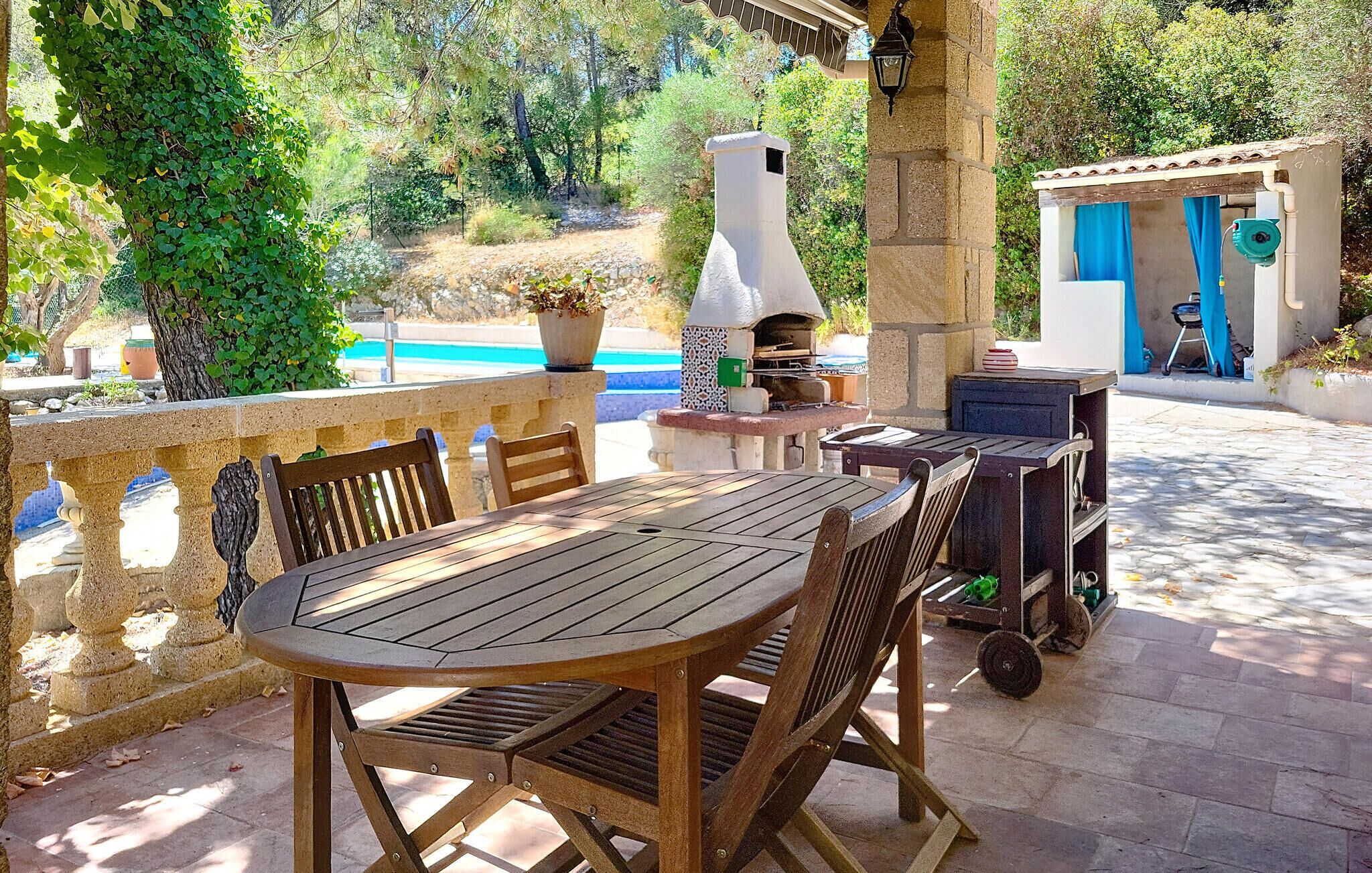 An outdoor patio with a round wooden table and chairs, a stone balustrade, a stone chimney, and a small building with a blue door.